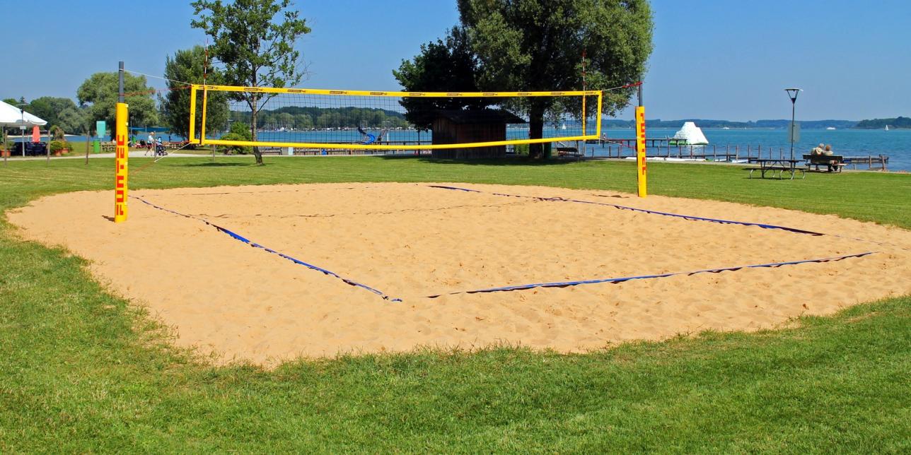 Cancha de voleibol al aire libre, con una superficie de arena suave en la zona de juego y rodeada de césped verde. La red está colocada en el centro, dividiendo el campo en dos mitades simétricas. El entorno natural crea un ambiente relajado, ideal para actividades deportivas al aire libre.