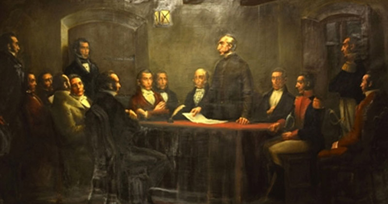 Grupo de hombres con vestimenta de época alrededor de una mesa. Algunos de ellos están de pie y otros sentados. En el centro un sacerdote sostiene un papel y sobre la mesa hay una pluma