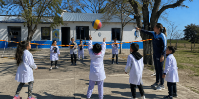 Grupo de niños y niñas con túnica blanca jugando vóleibol con una pelota colorida en el patio de una escuela rural; una docente con ropa deportiva señala la red mientras acompaña la actividad.