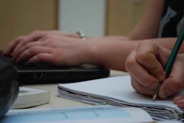Imagen que muestra las manos de dos personas adultas en actitud de aprendizaje (escribiendo sobre un papel y apoyándose sobre un ordenador)
