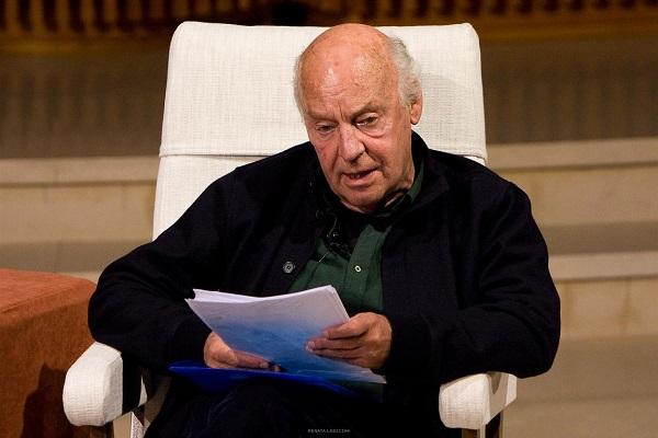 Fotografía de Eduardo Galeano en plano medio, sentado en un sofá y con unas hojas en su mano.