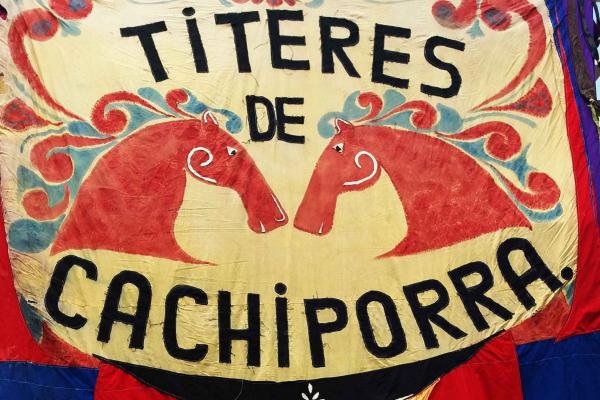 Bandera en tonos rojos y azules con las imágenes de las cabezas de dos caballos y el nombre de la compañía titiritera: "Títeres de Cachiporra" 