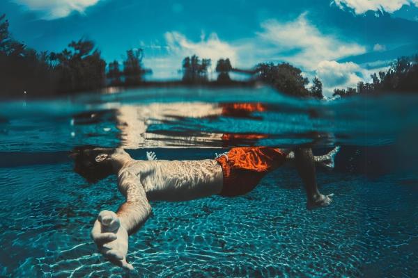 Hombre flotando en el agua, con los brazos extendidos. Cielo celeste con algunas nubes. 