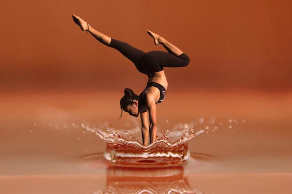 Mujer realizando una parada de manos sobre una superficie que simula agua, con un fondo degradado en tonos marrones. La postura refleja equilibrio y fuerza, mientras el agua alrededor forma un efecto visual de salpicadura.