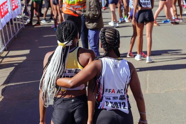 Dos mujeres atletas, vistas de espaldas, caminan abrazadas después de una carrera en la vía pública. Llevan ropa deportiva. Alrededor se observan otras corredoras y personal del evento en la línea de llegada.