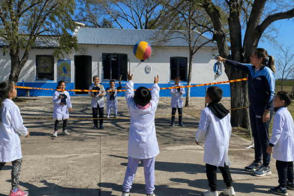 Grupo de niños y niñas con túnica blanca jugando vóleibol con una pelota colorida en el patio de una escuela rural; una docente con ropa deportiva señala la red mientras acompaña la actividad.