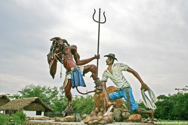 Escultura al aire libre que representa el enfrentamiento entre un diablo y un herrero. El diablo, de piel roja, cuernos y tridente, se muestra desafiante frente al hombre, que viste camisa blanca, pantalón azul y sombrero negro, sosteniendo un machete. Ambos están sobre una base rocosa, con vegetación y casas de techo de paja al fondo, bajo un cielo nublado.