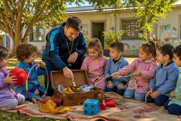 Docente arrodillado en el patio de una escuela abre una valija de madera ante un grupo de niños y niñas de 4 y 5 años sentados en círculo sobre una manta; alrededor hay objetos de juego como una pelota roja, un aro, una cuerda, un dado de tela azul y un silbato.