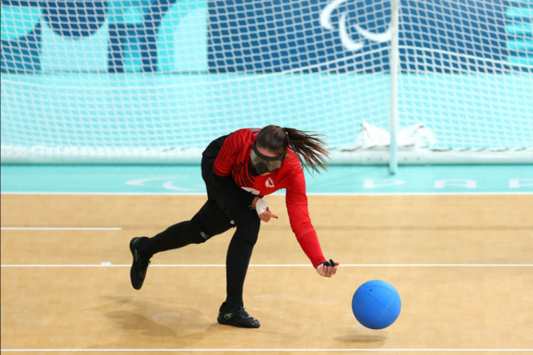 En la imagen se observa a un deportista con discapacidad visual en plena acción sobre la cancha de Goalball. Viste una camiseta deportiva roja de manga larga y pantalón deportivo largor y utiliza antiparras negras totalmente opacas que garantizan la igualdad de condiciones. Se encuentra próximo al contacto con la pelota azul con cascabeles caraterística de este deporte