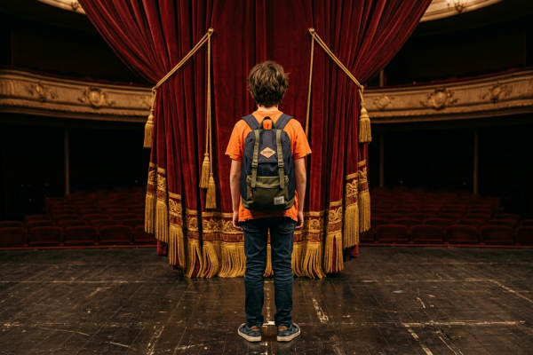 Niño vestido con remera de color naranja y jeans con una mochila, parado en un escenario frente al telón de un Teatro.