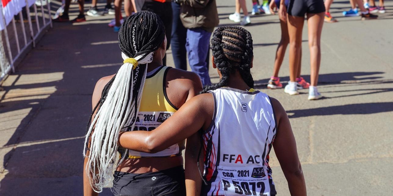 Dos mujeres atletas, vistas de espaldas, caminan abrazadas después de una carrera en la vía pública. Llevan ropa deportiva. Alrededor se observan otras corredoras y personal del evento en la línea de llegada.
