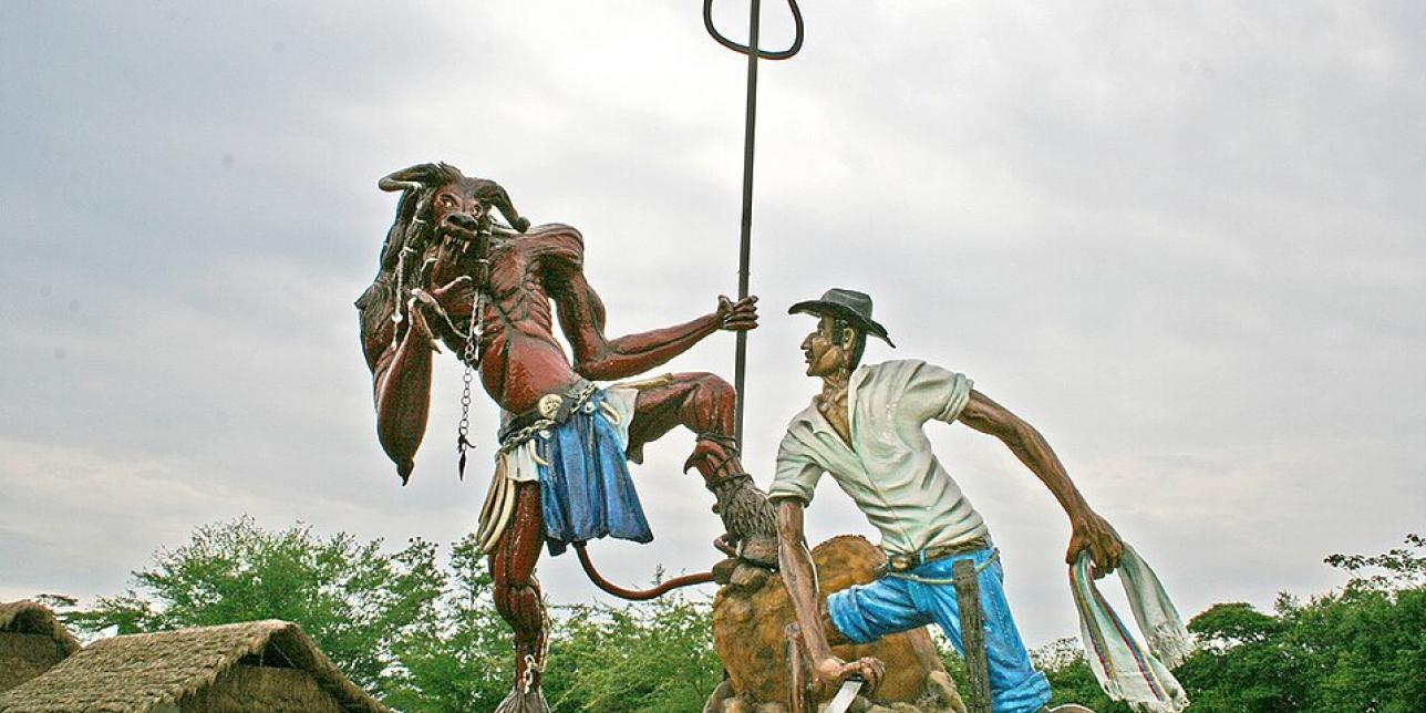 Escultura al aire libre que representa el enfrentamiento entre un diablo y un herrero. El diablo, de piel roja, cuernos y tridente, se muestra desafiante frente al hombre, que viste camisa blanca, pantalón azul y sombrero negro, sosteniendo un machete. Ambos están sobre una base rocosa, con vegetación y casas de techo de paja al fondo, bajo un cielo nublado.