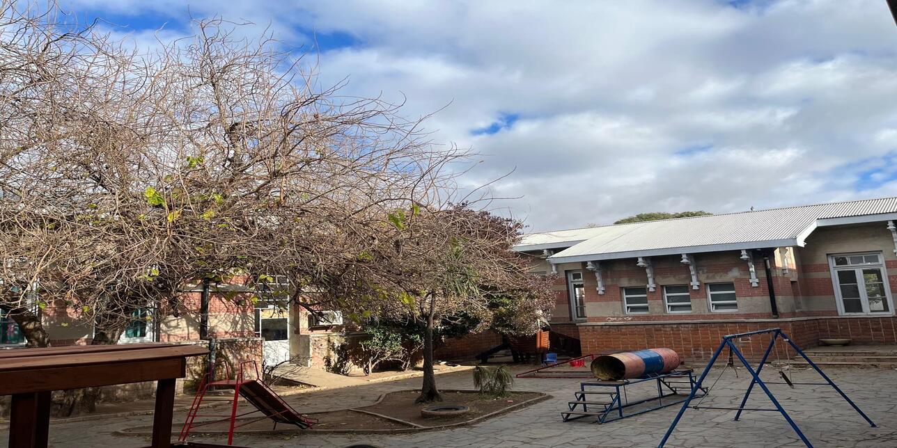 Patio escolar en invierno. Un árbol grande y otro más pequeño con ramas aparentemente secas. Un juego para pasar por un tanque encima de escaleras. Un tobogán y hamacas. Alrededor se visualiza parte del edificio escolar.